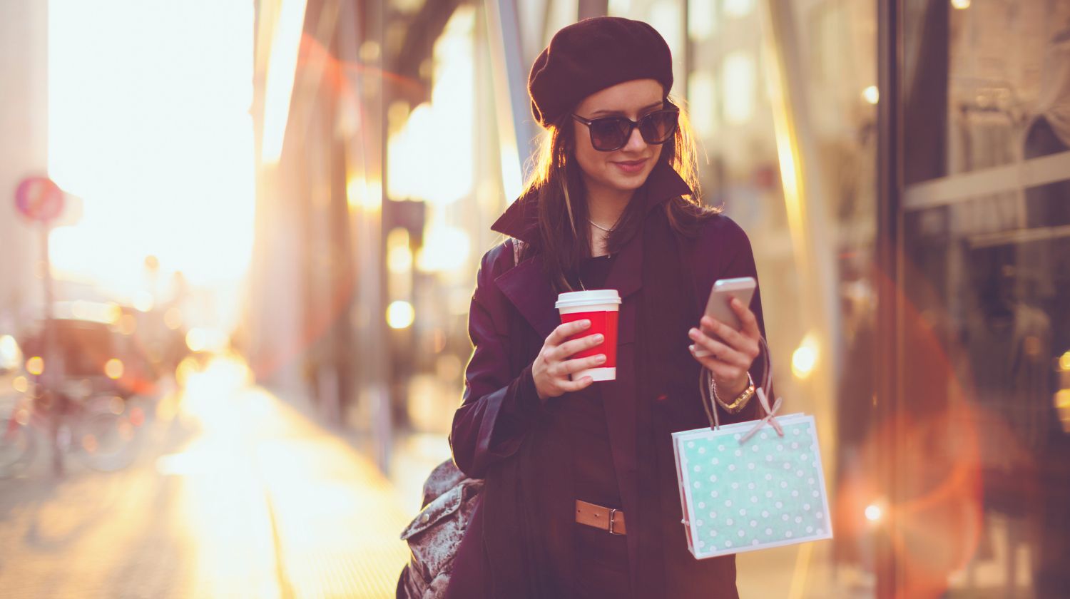 Žena stoji na ulici sa mobilnim i kafom u rukama